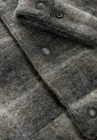 Grau- und braunfilzene Wollstoffe mit weicher Textur, versehen mit Metallschnappverschlüssen und Nähedetails. Mit horizontalen Streifen gemustert.