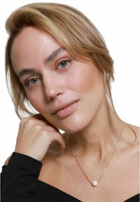 Delikat gullkjede med en sentral rund hvit perle. Modell kledd i en svart off-shoulder topp, lett posert med hånden på haken.