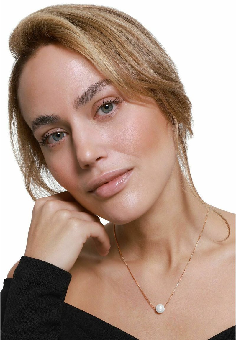 Delikat gullkjede med en sentral rund hvit perle. Modell kledd i en svart off-shoulder topp, lett posert med hånden på haken.