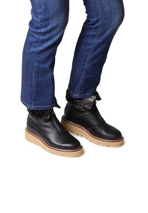 Bottes de cheville en cuir noir avec doublure en faux fourrure, fermeture éclaire et semelle en caoutchouc beige. Portées avec un jean bleu. Les bottes ont un design à bout rond.