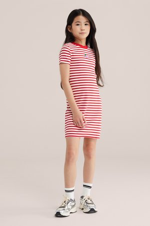 Jauna meitene stāv ģērbusies sarkanbalti svītrainā kleitā ar mazu zemeņu rakstu, baltās zeķēs un pelēkos sporta apavos.