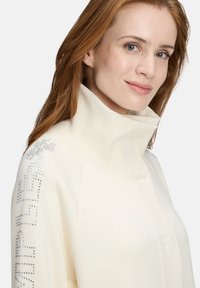 Sweat-shirt crème avec un col haut en côtes, orné de paillettes brillantes le long de l'épaule. Texture douce et lisse.