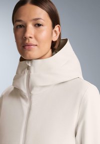 Svijetlosmeđi sako s visokim ovratnikom i prednjim patentnim zatvaračem. Materijal izgleda glatko s minimalnim šavovima i čistim dizajnom.