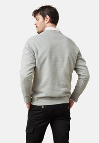 Sudadera gris claro hecha de una tela suave, con un escote redondo y puños acanalados. El modelo lleva pantalones negros con botones en el costado.