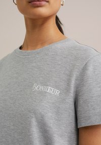 Siva bombažna majica s krožnim izrezom, z diskretno belo vezenino "BONHEUR" na levi strani prsnega koša, gladka tekstura.