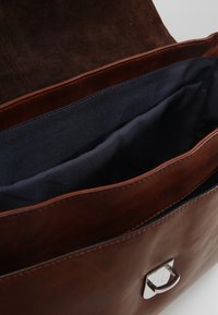 Borsa in pelle marrone con chiusura a patta, dotata di un accento hardware quadrato in argento. L'interno è foderato in tessuto blu e presenta diversi scomparti.
