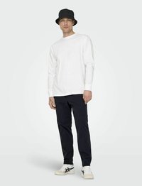 Witte katoenen shirt met lange mouwen, gecombineerd met marineblauwe broek, zwarte bucket hat en witte sneakers met zwarte accenten. Minimalistisch ontwerp.