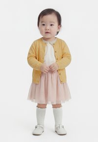 Väikelaps, kes kannab kollast kardigani, valget särk ja roosat tuultega seelikut, valget põlveni sokid ja valgeid kingi, seisab valgel taustal.