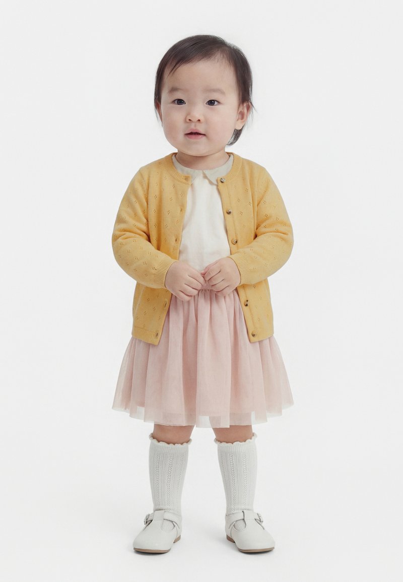 Väikelaps, kes kannab kollast kardigani, valget särk ja roosat tuultega seelikut, valget põlveni sokid ja valgeid kingi, seisab valgel taustal.