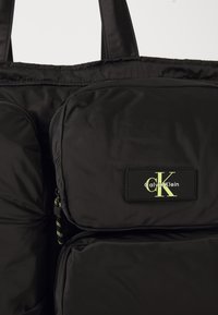 Black fabric bag with multiple zippered compartments, featuring a Calvin Klein logo patch in black and yellow on the front pocket.