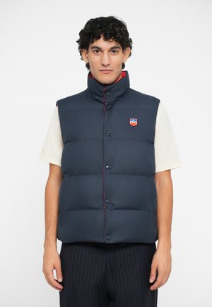 Navy blue puffer vest with high collar, red stitching, front buttons, and logo patch. Worn over a cream shirt, paired with pinstripe pants.