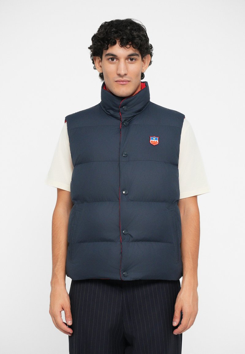Navy blue puffer vest with high collar, red stitching, front buttons, and logo patch. Worn over a cream shirt, paired with pinstripe pants.