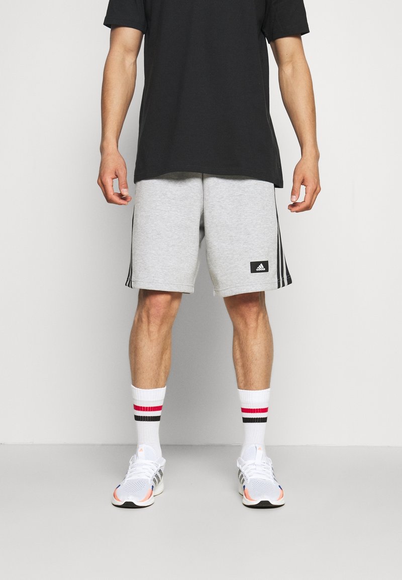 Pantalones cortos deportivos grises con un logo negro de Adidas, combinados con una camiseta negra y calcetines blancos con franjas rojas y negras. Zapatillas de running blancas.