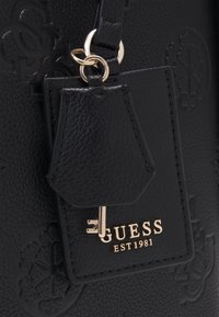 Borsa a tracolla in pelle nera con texture, caratterizzata da un logo dorato "GUESS EST 1981" e un ciondolo a forma di chiave attaccato a un'etichetta. Accentuata da un design floreale.
