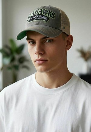 Junger Mann mit heller Haut, der eine graue Seattle Seahawks Mütze und ein weißes T-Shirt mit Rundhalsausschnitt trägt, blickt mit neutralem Gesichtsausdruck geradeaus.