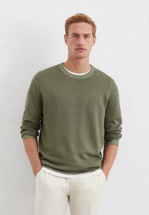 Olijfgroene sweater met een ronde hals en ribgebreide manchetten, gedragen over een wit shirt, gecombineerd met crèmekleurige broek.