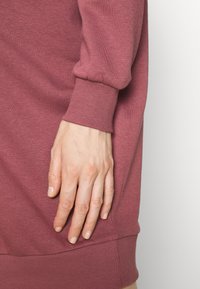 Una mano femminile poggiata sull'anca, che indossa un maglione a maniche lunghe con polsini a coste di colore rosa polveroso, su uno sfondo bianco semplice.