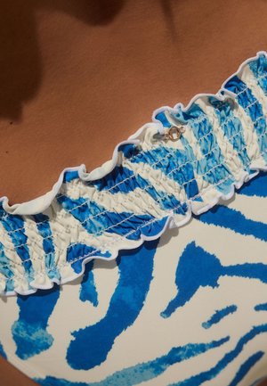 Close-up van blauw-wit gestreept badpaktopje met ruches en metalen gesp detail op gebruinde huid.