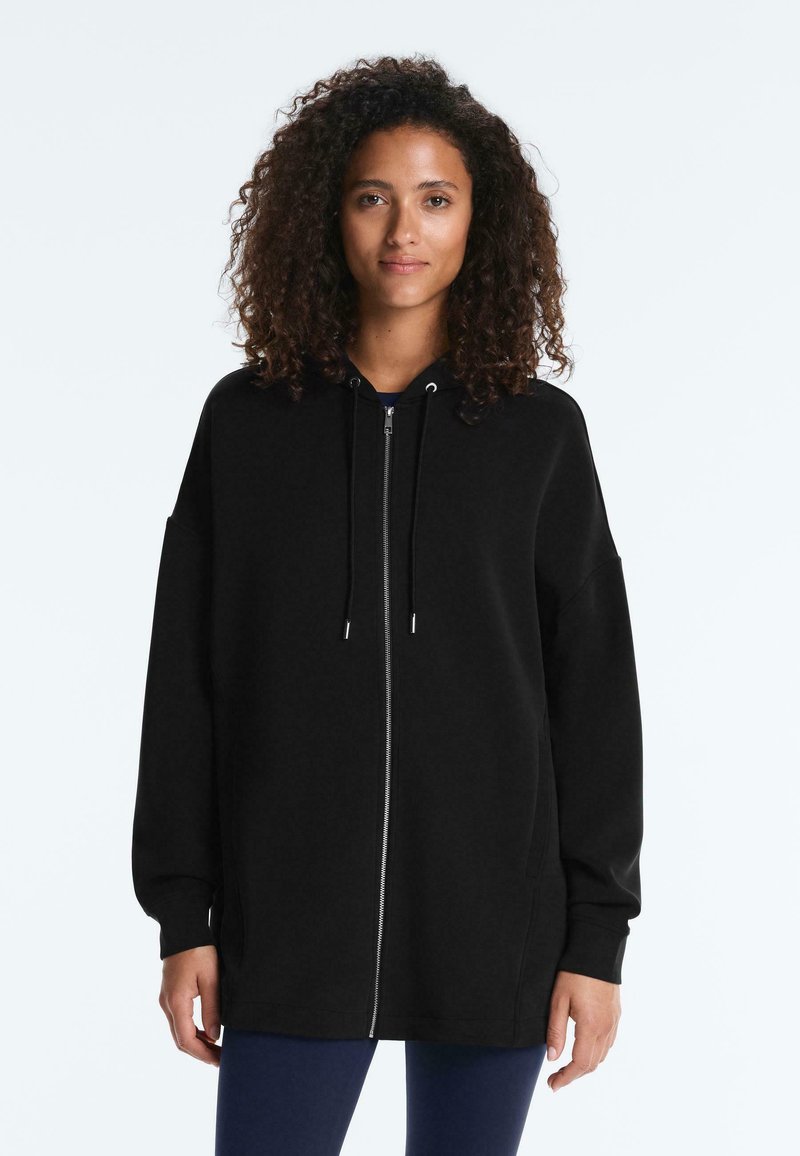 Femme aux cheveux bouclés portant un long sweat à capuche noir zippé et des leggings bleu marine, debout devant un fond blanc uni.