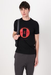 Juodas medvilnės marškinėliai su ryškiu raudonu ir juodu grafiniu logotipu, esantys ant krūtinės. Modelis laiko tamsią pečių rankinę, vilkėdamas pilkas kelnes.