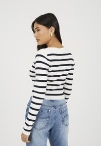 Pasiasty sweter z długim rękawem w kolorze białym i granatowym. Żebrowana tekstura, dopasowany fason, okrągły dekolt, noszony z jasnoniebieskimi spodniami o wysokim stanie.