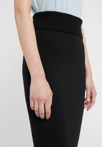 Gros plan sur le bas du torse d'une personne portant une jupe tricotée noire taille haute et un haut bleu clair, les mains détendues sur les côtés.