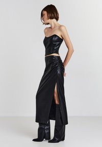 Femme en top sans manches en cuir noir et jupe fendue, associée à des bottes hautes assorties, se tenant de côté devant un fond uni.