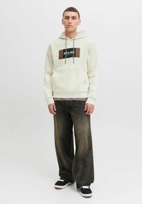 Felpa crema con un logo rettangolare nero e marrone, abbinata a jeans larghi scuri e sneaker nere con suole bianche.
