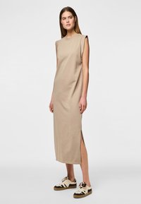 Vestido beige sin mangas hecho de una tela suave, con aberturas laterales y un corte relajado, combinado con zapatillas blancas y negras.