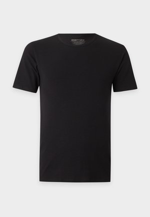 Mann steht barfuß und trägt ein schwarzes T-Shirt mit kurzen Ärmeln und schwarze Boxershorts vor einem schlichten weißen Hintergrund.