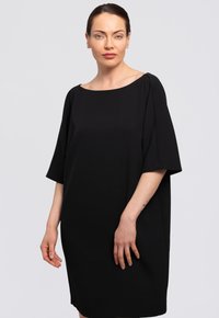 Musta mekko, jossa on löysä, lyhythihainen muotoilu ja leveä kaula-aukko. Valmistettu sileästä kankaasta, se ulottuu polven ylle suoralla siluetilla.