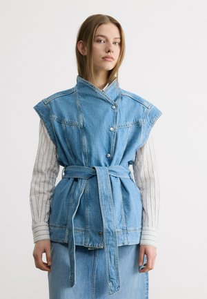 Mladá žena v bezrukávové džínové vestě s páskem přes proužkované tričko s dlouhým rukávem a džínovou sukní, stojící před jednobarevným pozadím.