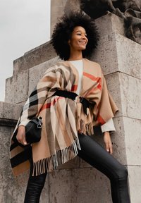 Poncho à carreaux beige avec des rayures noires, brunes et rouges, présentant des bords frangés. Porté par-dessus un pull blanc et associé à des leggings noirs.