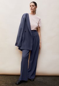 Marineblauwe jas over een lichtroze T-shirt, gecombineerd met bijpassende marineblauwe broek en zwarte schoenen. Soepele textuur, op maat gemaakt.