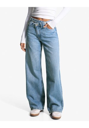 Person iført lyseblå brede jeans, hvid crop top med lange ærmer og hvide sneakers med gummisåler, stående med den ene hånd i lommen.