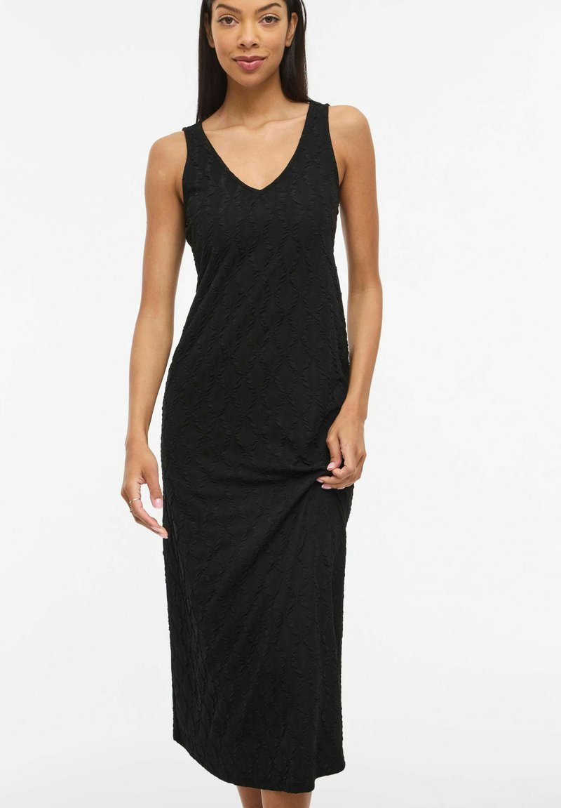 Femme aux longs cheveux foncés portant une robe midi noire sans manches à texture avec un col en V, posant devant un fond blanc uni.