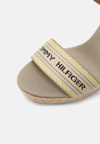 Beige espadrille sandal med en bred stofrem, der har sorte og gule striber, præget tekst og en vævet jute-sål.