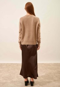 Beige gebreide trui met een ribbelsrand, gedragen over een bruine satijnen midi-rok, gecombineerd met zwarte chunky hakken. Soepele textuur, relaxte ontwerp.