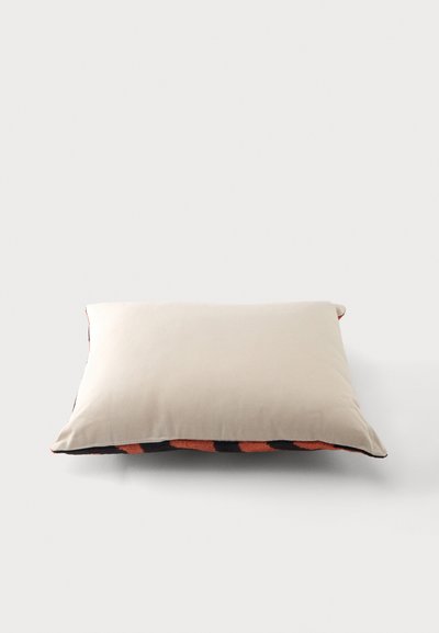 Coussin rectangulaire beige avec un bord visible montrant un tissu à motifs noirs et orange en dessous sur un fond blanc.