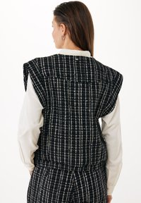 Černobílý kostkovaný vest, vyrobený z texturované tkaniny, s širokými rameny, otevřeným zadním dílem a vrstvenou bílou blůzou pod ním.