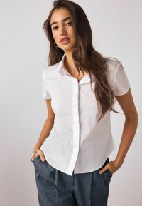 Biela košeľa s krátkym rukávom a golierom, s gombíkmi na prednej strane a priliehavým strihom. Jednofarebná látka bez vzorov.