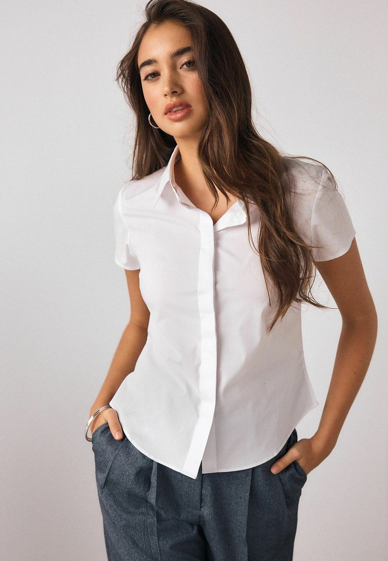 Biela košeľa s krátkym rukávom a golierom, s gombíkmi na prednej strane a priliehavým strihom. Jednofarebná látka bez vzorov.