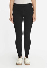 Schwarze Leggings aus dehnbarem Stoff, mit hohem Bund und enganliegendem Schnitt. Kombiniert mit weißen und beigen Turnschuhen.