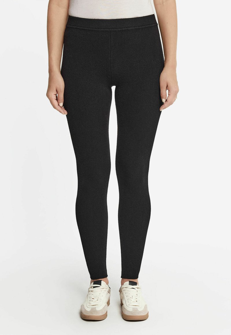 Schwarze Leggings aus dehnbarem Stoff, mit hohem Bund und enganliegendem Schnitt. Kombiniert mit weißen und beigen Turnschuhen.