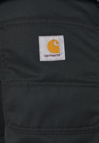 Svart tyg med en diagonal textur. En fyrkantig vit tagg har en gul logotyp och texten "carhartt", placerad på en ficka.