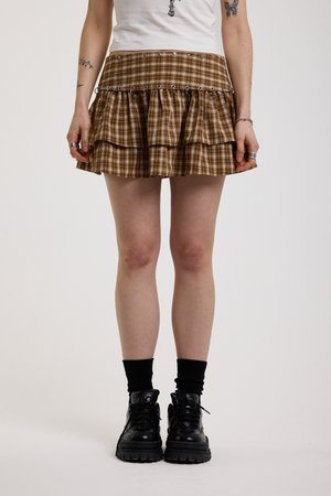 Άτομο που φοράει καφέ μίνι φούστα σε στρώσεις με μεταλλικά κουμπώματα, μαύρες κάλτσες, χοντρά μαύρα παπούτσια και λευκό τοπ χωρίς μανίκια με τατουάζ στα χέρια.