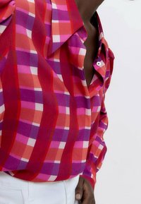 Camicia button-up a quadri rossa e rosa con colletto, caratterizzata da tonalità diverse di rosa e viola in un tessuto morbido. I bottoni bianchi aggiungono dettagli.