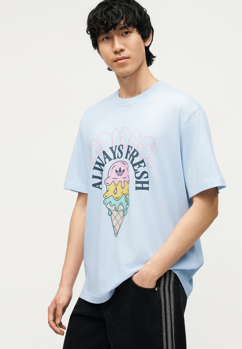 Joven con pelo negro y rizado que lleva una camiseta azul claro con un cono de helado colorido derritiéndose y el texto “ALWAYS FRESH”.