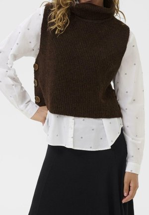 Kobieta w brązowej, dzianinowej kamizelce bez rękawów z bocznymi guzikami, na białej koszuli zapinanej na guziki i czarnej spódnicy, z ręką na biodrze.