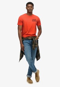 Camiseta roja de algodón con un logo, jeans azules y botas de ante color crema. Camisa a cuadros verdes atada a la cintura. Ajuste casual, mangas cortas.